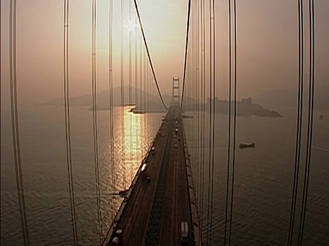 夕阳落在桥后，汽车在桥上行驶，太阳反射金色的光在下面的河流和朦胧的风景在B/G。LA, MS光束在桥结构顶部对天空，当摄像机转动光束移动PAN右，沿倾斜下来视频素材