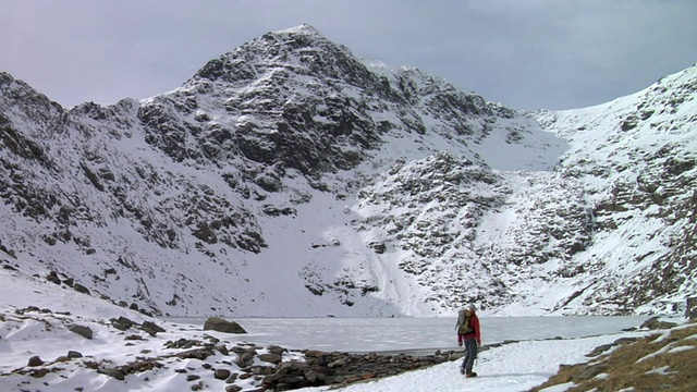 登山者走在冰冻的山湖，背景是白雪覆盖的山/ Llanberis，斯诺登尼亚，英国视频素材