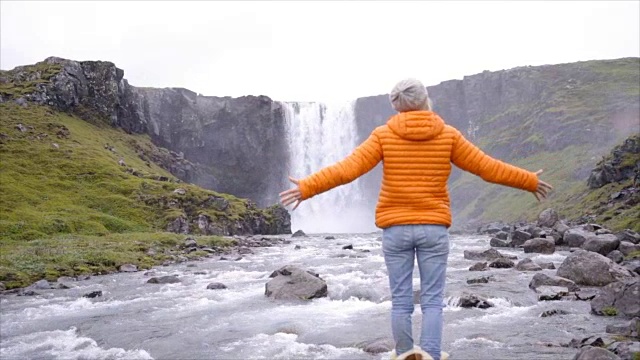 一名年轻女子在冰岛壮丽的瀑布前张开双臂。人们旅行探险的概念——慢动作视频素材