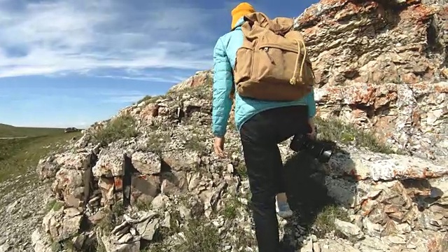 就像一个从后面戴着帽子去当摄影师的女孩。一个旅游摄影师背着背包，手里拿着相机，正沿着岩石高原走向悬崖峭壁，欣赏风景。慢镜头4 k视频素材