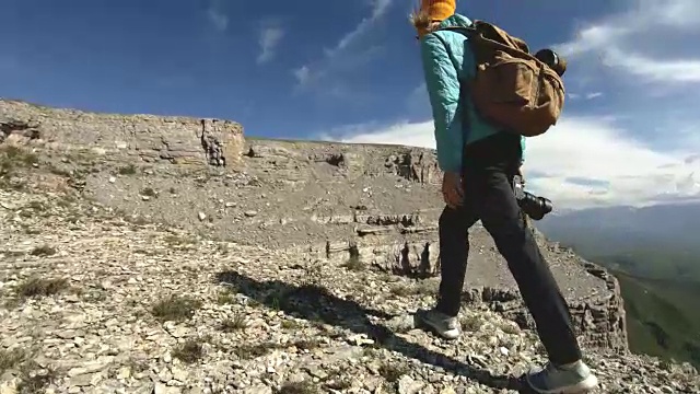 就像一个从后面戴着帽子去当摄影师的女孩。一个旅游摄影师背着背包，手里拿着相机，正沿着岩石高原走向悬崖峭壁，欣赏风景。慢镜头4 k视频素材