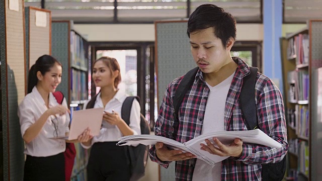 年轻快乐的亚洲学生男人在大学图书馆看书和她的同学在背景，教育和学校的理念视频素材