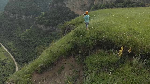 戴着眼镜和帽子的女孩摄影师在悬崖附近的高原边缘用她的单反相机拍了一张照片。鸟瞰图视频素材