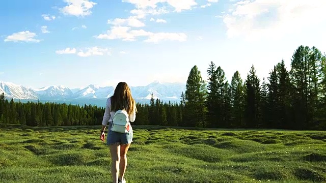 一个背着背包的女孩在森林和白雪皑皑的山脉上绕着田野走。视频下载