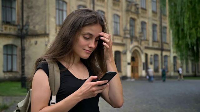 年轻漂亮的白人学生用手机打字，站在大学附近的公园里，女人拿着背包视频素材