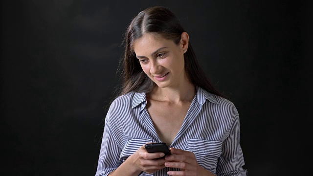 漂亮的年轻女子穿着条纹衬衫发短信，微笑着，孤立地站在黑色背景视频素材