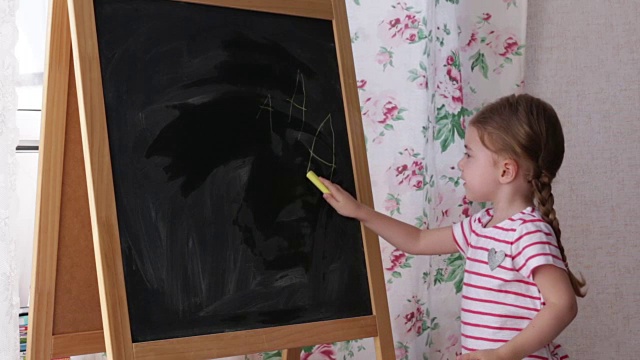 学龄前小女孩在黑板上写字。蹒跚学步的小女孩拿着粉笔画画视频下载