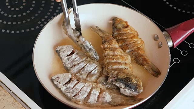 人在平底锅里用油炸鱼。特写男子的手flips鱼与烹饪钳。视频素材