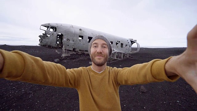 4k-一名年轻人站在黑沙滩上的飞机残骸旁自拍。这是冰岛著名的旅游景点，并与飞机残骸合影视频素材