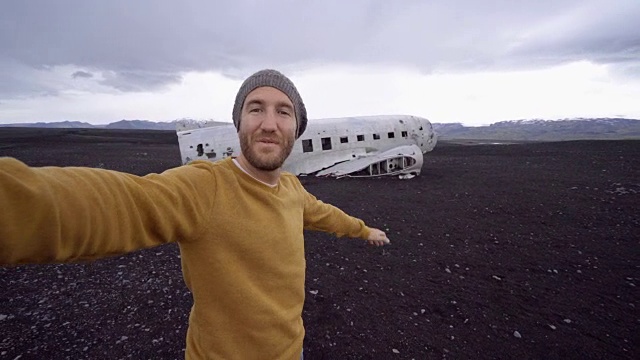 4k-一名年轻人站在黑沙滩上的飞机残骸旁自拍。这是冰岛著名的旅游景点，并与飞机残骸合影视频素材