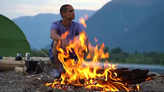 一名游客坐在山湖边的火堆旁。视频素材