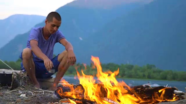 一名游客坐在山湖边的火堆旁。视频素材