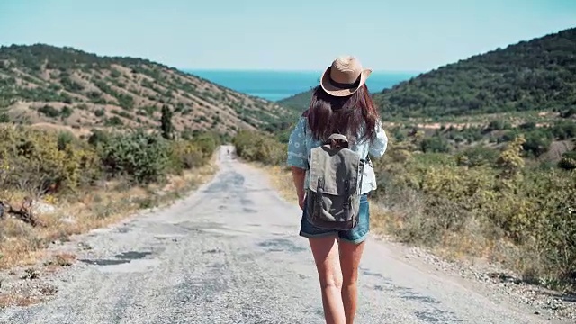 后视图活跃的背包女戴着帽子走在通往大海的道路上，周围群山环绕视频素材