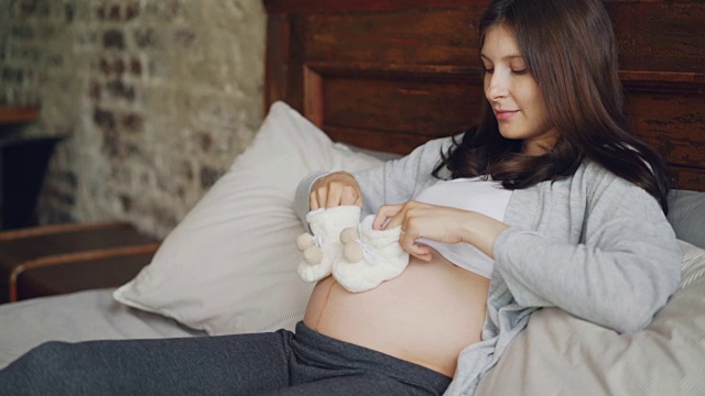 怀孕的年轻女士手里拿着可爱的婴儿鞋，在她的肚子上迈着小步，玩着未出生的孩子，用爱看着她的肚子。视频素材