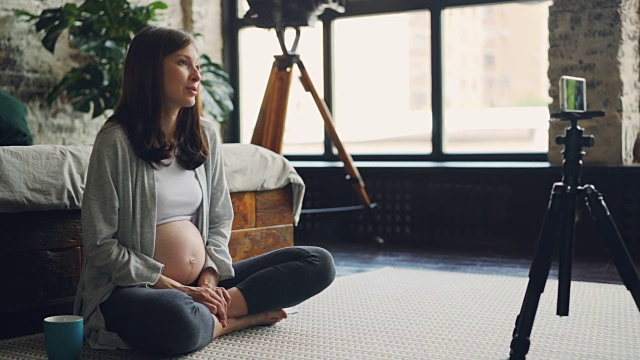 期待母亲的女博主正在为网络博客录制关于健康怀孕的视频，女孩正看着三脚架上的智能手机摄像头说话。视频素材