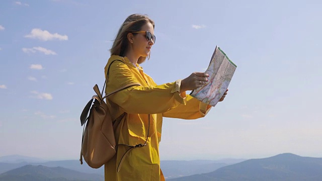 一名穿着雨衣的年轻女子在山间小径上徒步旅行，停下来查看地图，寻找方向。徒步者到达山顶，查看地图上的方向视频素材