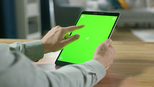 一名男子在绿色模拟屏幕的数字平板电脑上用手势肖像模式坐在他的办公桌前。在舒适的客厅背景。视频素材