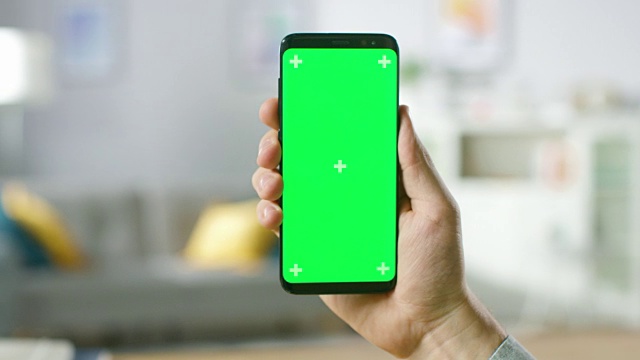 一个男人的手握绿色模拟屏幕智能手机的特写。现代移动电话。在舒适的客厅或家庭办公室的背景。视频素材