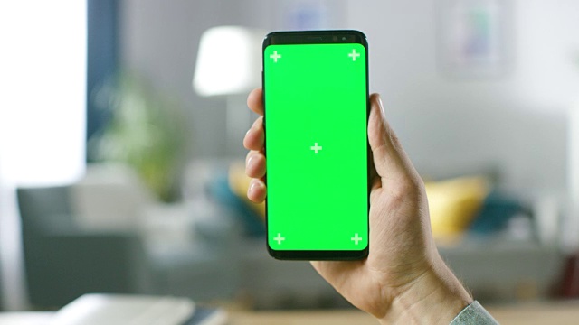 一个男人的手握现代智能手机与绿色模拟屏幕的特写。在舒适的客厅或家庭办公室的背景。视频素材