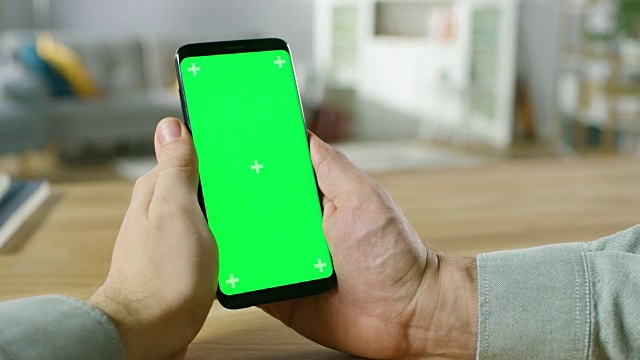 一个人拿着现代绿色模拟屏幕智能手机在他的手中的特写。使用现代高端设备。在舒适的客厅或家庭办公室的背景。视频素材