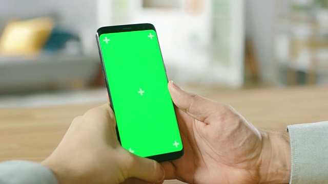 一个人拿着绿色模拟屏幕智能手机在他的手中的特写。使用无线设备上网浏览。在舒适的客厅或家庭办公室的背景。视频素材