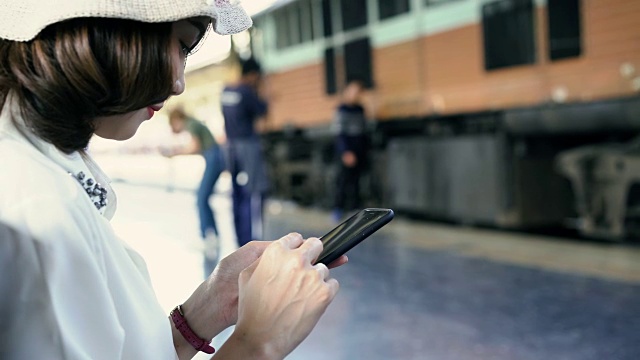 旅行家背包亚洲女人旅行在曼谷，泰国。快乐的年轻女性坐在长凳上用智能手机聊天，阅读和发短信在火车站。视频素材