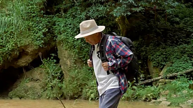 年长的亚洲人享受走在瀑布前。旅行的生活方式和成功的概念度假到野外穿着舒适的衬衫，帽子和背包视频素材