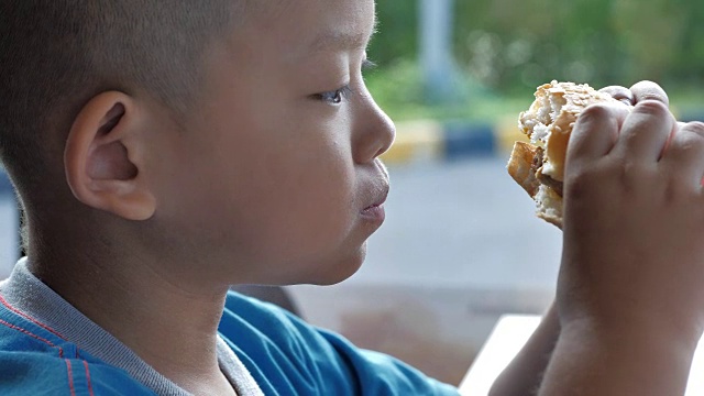 近距离的亚洲小男孩喜欢吃汉堡，可爱的快乐的男孩拿着汉堡在餐厅。视频慢动作视频素材