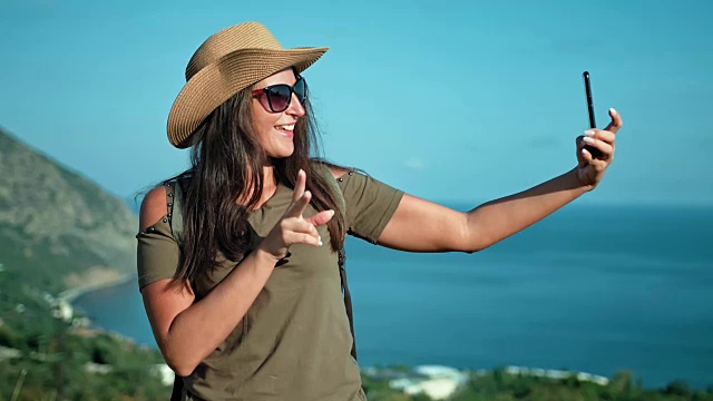 带着帽子和太阳镜的微笑女人旅行者的肖像，用智能手机摆姿势自拍视频素材