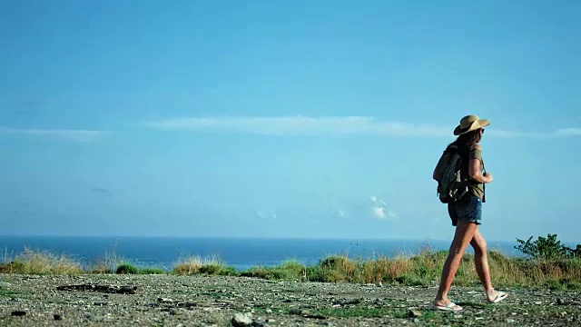 背包女徒步旅行者戴着太阳镜和帽子走在山的边缘，越过海边的景色视频素材