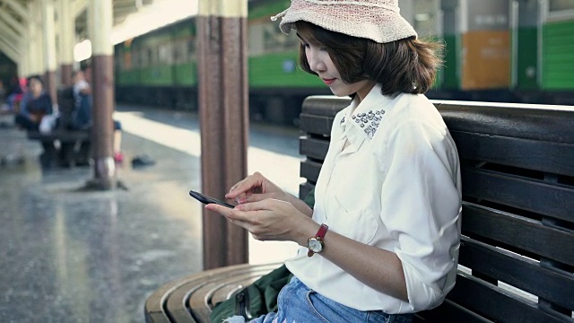 旅行家背包亚洲女人旅行在曼谷，泰国。快乐的年轻女性坐在长凳上用智能手机聊天，阅读和发短信在火车站。视频素材