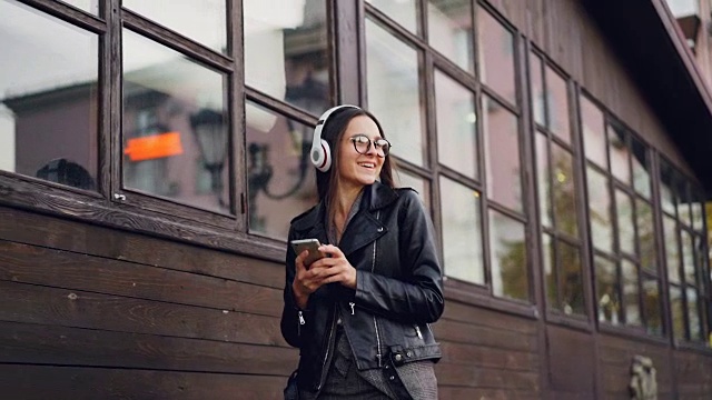 迷人的女孩通过耳机拿着智能手机听音乐，独自走在街上玩的慢动作。青年与科技的概念。视频素材