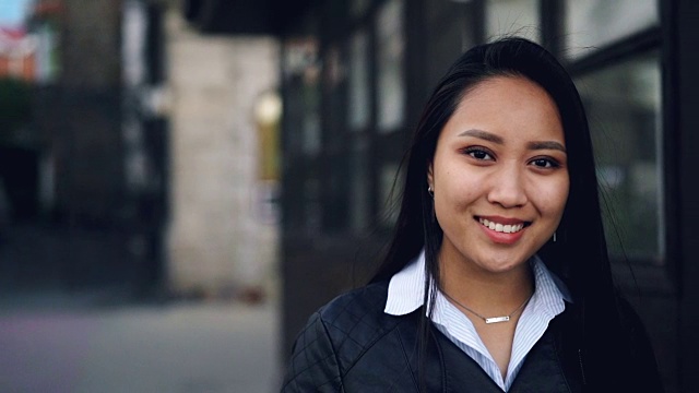 特写慢动作肖像迷人的亚洲女孩看着镜头与幸福的微笑站在街上穿着时髦的衣服皮夹克和衬衫。视频素材