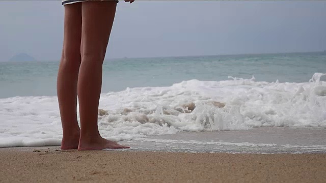 一个女人光着脚走在夏天的海滩上。年轻女子的腿在海滩上走在海浪和沙子。旅行的概念。视频素材