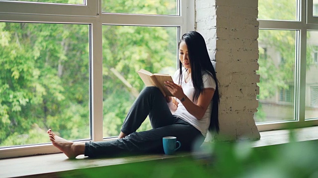 迷人的亚洲女学生正在阅读和微笑坐在窗台在现代公寓。爱好、青年文化及室内理念。视频素材