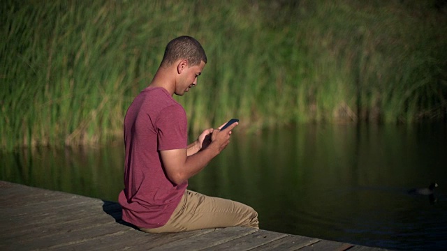 一名年轻男子坐在湖边的码头上发短信视频素材