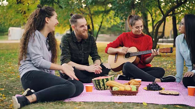 多民族的朋友们在公园里唱歌和拍手，而美丽的女孩在野餐时弹吉他。食物和毯子是可见的。视频素材