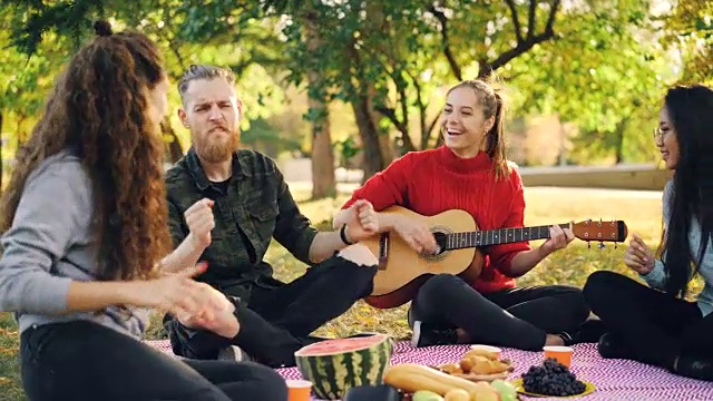 顽皮的年轻人唱歌和移动的手，当美丽的女孩弹着吉他在公园野餐在阳光明媚的秋天。乐趣和音乐的概念。视频素材