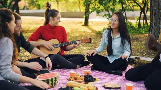 在阳光明媚的秋日野餐时，当他们的朋友漂亮的女孩在弹吉他时，兴奋的女孩和男孩在唱歌和拍手。树木，毯子和食物都看得见。视频素材