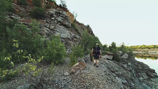 一个游客背着背包沿着湖岸走着，背景是高高的岩石和野生的自然视频素材