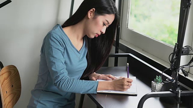 年轻漂亮的亚洲女人在桌子上的笔记本上写字，女孩在家里用书或日记工作，女孩是作家，自由商业概念，慢动作。视频素材