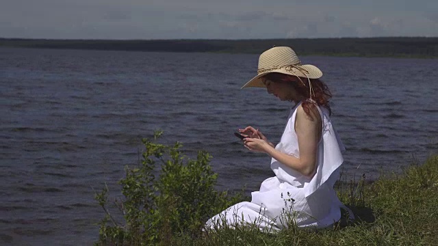 美丽的红发女孩穿着白色的夏装坐在岸边和河边玩手机视频素材