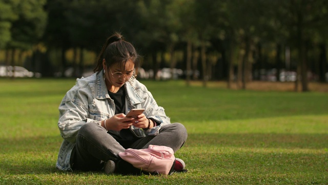 亚洲大学女孩坐在草地上使用手机在校园里视频素材