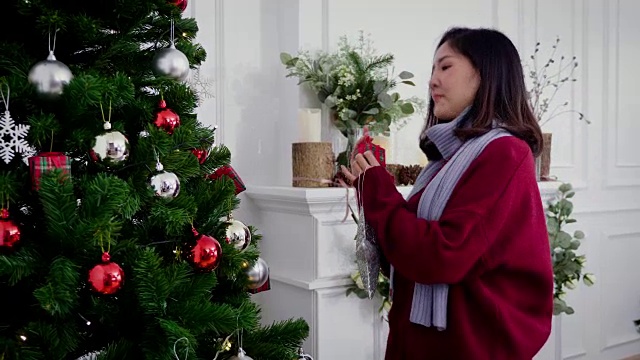 年轻的亚洲女人在毛衣装饰圣诞树与装饰品在客厅在家里，冬季假期和圣诞节的概念视频素材