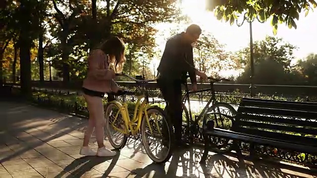 一对美丽的年轻夫妇在空旷的城市公园里骑着自行车。停下来，坐在长椅上拥抱。爱的夫妻。阳光照在背景上。加速视频视频素材