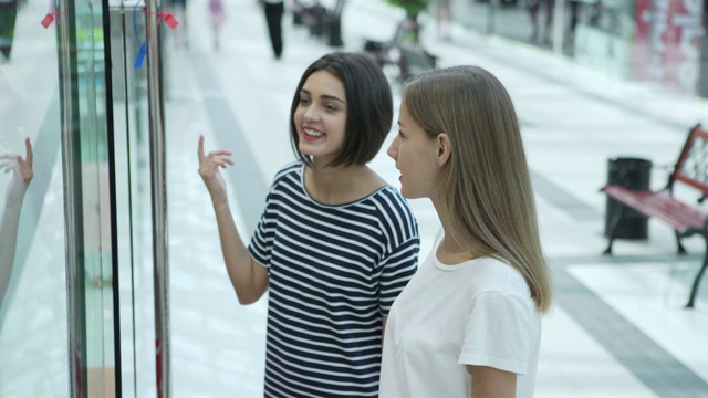 两个兴高采烈的女孩站在购物中心的橱窗前讨论新系列的衣服视频素材