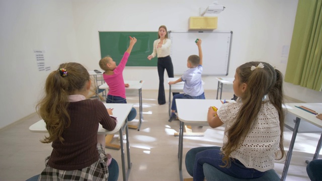 幼儿学习时，微笑的教师在黑板旁为小学生在学校的课桌上进行认知课视频素材