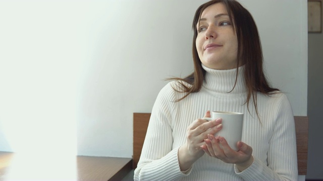 穿着白毛衣的年轻女子在咖啡馆喝咖啡。视频素材