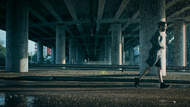 一个雨天，一个穿着运动服的肌肉发达的年轻人在街上慢跑。他在桥下的城市环境中跑步。视频素材