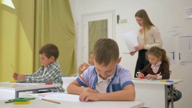 小学的孩子们用彩色铅笔在纸上和年轻的老师在灯光教室视频素材